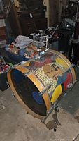 Photo showing side view of vintage Tama bass drum with colorful hand painted shell and attached metal hardware, drum is placed on floor in storage area.