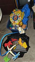 Photo showing a plastic storage bin filled with various vintage baby toys including a circular animal-themed toy and a Fisher Price mobile with blue plastic parts, next to a black bag containing parts of a toy train set.