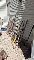 Photo showing a group of assorted yard tools leaning against a wall outdoors on a paved surface. Tools include various shovels, rakes, a yellow paint roller extension, pick, and stirrers. All show signs of use and dirt.
