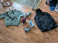 Photo shows two jackets (green with 'ARMY' lettering and black jacket with orange logo), a red handheld vacuum with charger, the repair manual, and indoor door knob packaging on a floor.