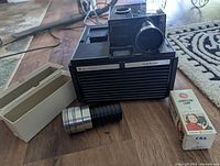 Bell & Howell Slide Cube Projector Model 977Q with detached cylindrical zoom lens and quartz lamp box on hardwood floor.