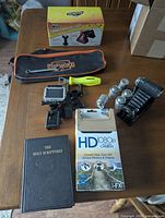 Overall photo showing the set of items on a table including the vintage folding camera, film tins, HD 1080p camera with accessories, Jewish Bible, tool bag, car care product box and Snap-on bottle opener.