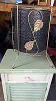 Front view of wire wastepaper basket sitting on the cabinet, showing black perforated metal basket and decorative gold leaf frame
