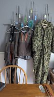 Three hanging garments: green camo one-piece suit, brown jacket, and brown pants with suspenders, displayed on hangers against wall hooks.