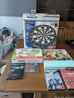 Hannex Alpha dartboard boxed, Rugrats Desktop Pinball, 1000 Masterworks puzzle, 50 Cent CD, Playstation 2 game, and DVDs on table