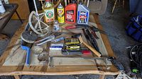 Photo showing a variety of garage tools and fluids on a wooden table including hand saw, wire brush, spark plugs, lubricant bottles, and hand tools.