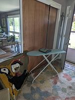 Ironing board set up with woven picture leaning beside it and rug sweeper resting on the ironing board
