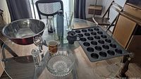 Photo showing all items together: large stainless steel bowl, crystal glass bowl, red aluminum travel mug, set of clear drinking glasses, 24-cup cupcake pan, salt or pepper shaker, and cups.
