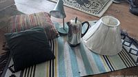 Photo showing two pillows, a Mid-Century Modern gooseneck lamp, a stainless steel percolator coffee pot, a new white lampshade and the rug beneath.