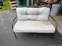 Outdoor iron bench with cushions on a paved area. Cushions are light beige/gray and have some visible dirt and wear.