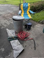 Full lot overview showing metal garbage can with lid, blue and yellow folding chair with cup holder, red extension cord, fabric landscaping material, portable sprinkler, small cup, and cooler.