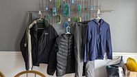 Photo showing all five clothing items hanging on a metal grid against a wall: Nautica jacket, black puffer jacket, gray ski pants, blue hoodie, and camo belt.