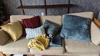 View of multiple pillows and pillow covers placed on a beige couch, showing various colors and textures including gold, blue chenille, burgundy, light grey, and dark grey pillows.