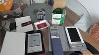 Photo showing multiple items including Literati eReader, Sony eBook reader, calculator, green bulb box, pen holder, and cords on a glass table.