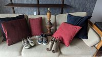 Couch with five pillows in dark blue, red, and maroon; three pairs of women shoes - beige ankle boots, navy sneakers, and green sandals; vintage wood lamp behind couch.