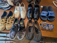 Seven pairs of shoes plus blue sports goggles displayed on wooden surface. Shoes vary in style, color, and condition.