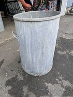 Full view of the galvanized metal garbage/burning can standing upright outdoors on pavement.