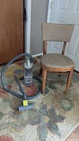 Photo of beige bentwood framed upholstered chair next to a compact hand-held vacuum cleaner with long hose and floor attachment.