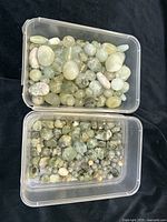 Two clear plastic containers filled with polished oval and round semi-opaque pale green and beige agate and quartz beads with natural inclusions.
