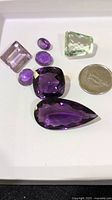 Seven faceted gemstones on white surface including six purple amethysts and one green amethyst, 10 cent coin for scale