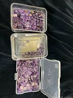 Wide view of three plastic containers holding loose amethyst beads of various cuts and shapes, primarily purple hues.