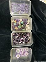 Four plastic containers with different assortments of gemstones including polished lavender chalcedony beads, rough mixed gemstones, tumbled amethyst, and amethyst stalactite slices with a large oval pendant.