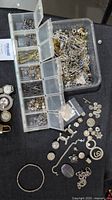 Wide view showing two plastic storage containers filled with assorted jewelry findings including clasps, pins, rings, and loose decorative parts on table.