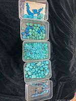 Five transparent plastic containers holding turquoise beads and stones of various shapes and colors arranged on a black surface.