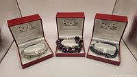Three bracelets displayed individually in red Amalia Zanetti boxes with white interior. Left: silver woven bracelet with crystal bands; center: dark black pearls double strand bracelet; right: triple strand crystal and pearl bracelet.
