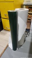 Two upright rolls side-by-side: white canvas roll and green glossy roll, both 24 inches wide