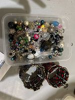 Plastic container filled with assorted costume jewellery beads in various styles and colors, alongside two silver-tone bracelets with red decorative stones.