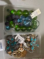 Photo shows two plastic containers with costume jewellery including beaded bracelets with green and blue beads, a silver chain with blue beads, and assorted earrings with turquoise stones and metal accents.