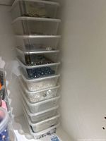 Stack of 11 transparent plastic bins filled with assorted crystal beads, various colors including blue, white, yellow, and clear beads, stored in a cabinet corner.