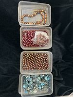 Four plastic containers stacked vertically showing freshwater pearl beads in brown, gold, burgundy, and blue. The top container has a strung pearl necklace with brown and gold pearls, the other containers contain loose pearls in their respective colors.