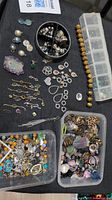 Photo showing wide variety of beads, charms and findings laid on black cloth including two plastic containers full of mixed beads.