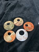 Five large polished stone disc pendants arranged on black fabric, showing variations in natural colors and patterns of agate and jasper.