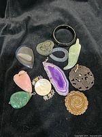 Ten various pendants and one black bracelet arranged on black velvet cloth, showing different gemstone types and carved details.