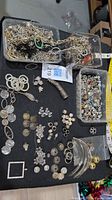 Photo showing two plastic containers filled with various silver colored metal chains, beads, and charms alongside laid out pendants and bead pieces on black surface.