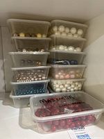 Plastic bins stacked in a corner filled with shell pearls and beads of various colors including white, cream, pink, blue, and red labeled Majorca