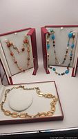 Three necklaces displayed in red display boxes: coral bead chain link necklace, turquoise and crystal bead necklace, and gold-plated chain with orange painted links