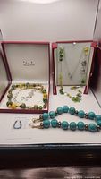 Three necklaces on display, two inside open jewelry boxes and one laid out in front. Green, yellow, brown African bead necklace and a silver chain necklace with green dyed jade beads present.