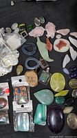 Wide view of various loose polished and cut gemstones as well as pendants lying on black cloth including leaf shapes, oval stones, and some packaged pieces.