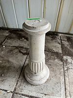 Front view of one concrete bird bath pedestal showing cylindrical shape with fluted midsection and rounded base on concrete floor with metal siding background.