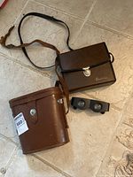 Two brown carrying cases and a pair of black binocular glasses on tiled floor.