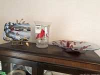 Three cardinal-themed decorative pieces displayed on a wooden cabinet: decorative plaque on a stand, glass vase with cardinal design, and a large round glass plate with cardinal painting and red berries.