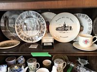 Various ceramic collectible plates and mugs displayed on wooden shelves, showcasing Canada Centennial 1867-1967 historic designs and provincial crests.