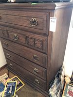 Full view of a tall wooden chest of drawers with five drawers, metal handles, and carved decorative panels on the second drawer down from the top.