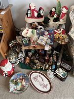 Wide view of assorted Christmas decorations including Santa and penguin figurines, mugs, plates, snow globe, stockings, small decorative houses, candles, and other holiday decor items displayed on shelves and floor.