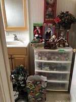 Photo showing 4-drawer plastic organizer filled with clear compartments of various Christmas ornaments and decorations, topped with boxed holiday figures including the Dancing Maxine Doll and Rockin' Rollers Snowman Skier figure, plus a poinsettia floral arrangement and other decor items.