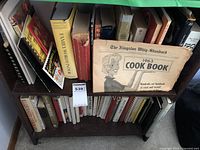 Wide view of shelf with assortment of cookbooks including the prominent vintage 1963 Kingston Whig-Standard Cook Book on top shelf